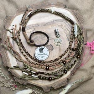 Könnte beinhalten: Handgefertigte Dreadlock-Extensions in Braun-, Grün- und Beigetönen, mit dekorativen Perlen und einem silbernen Anhänger. Enthält ein Haargummi und ein Logo "DreadArt by Annett."