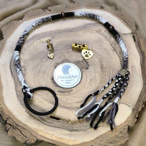 May include: A black, white, and grey dreadlock hair wrap with a black elastic band. The wrap includes decorative charms, such as a gold heart with the text "My Dog" and a paw print charm. The logo "DreadArt by Annett" is also visible.