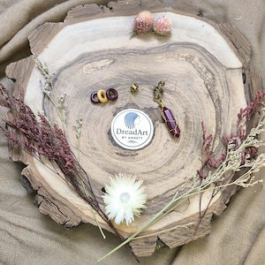 Könnte beinhalten: Eine Anordnung von Haarschmuck und dekorativen Elementen auf einer Holzscheibe. Die Artikel umfassen einen Anhänger mit einem weinroten Kristall, Haarperlen und einen Aufkleber mit dem Logo "DreadArt by Anett". Getrocknete Blumen und Blätter ergänzen das Arrangement.