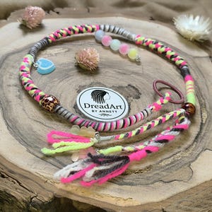 May include: A colorful dreadlock wrap featuring neon pink, yellow, and gray accents. The wrap includes decorative beads, a heart-shaped charm, and a logo that reads "DreadArt by Anett."