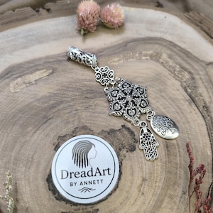 Puede incluir: Cuenta de rastas plateada con un diseño de filigrana ornamentado. La cuenta presenta un motivo floral central, un amuleto de mano de Hamsa y un amuleto de árbol de la vida. La cuenta se muestra sobre una superficie de madera con flores secas y un logotipo "DreadArt by Annett".