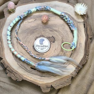 Op de afbeelding: Een handgemaakte dreadlock wrap in pastelkleuren, waaronder lichtgroen, blauw en perzik. De wrap heeft een gevlochten ontwerp en een lichtgroene elastische band. Het DreadArt-logo is zichtbaar op een witte sticker.