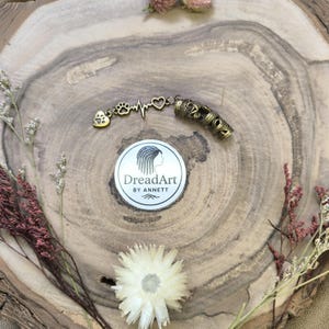 May include: A gold-toned dreadlock accessory featuring a paw print, heartbeat, and heart charm, connected to a patterned cylindrical piece. The image includes a sticker with the text "DreadArt by Annett".