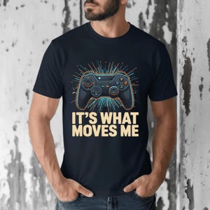 May include: Navy blue t-shirt featuring a stylized black video game controller design with orange and blue accents. The text "IT'S WHAT MOVES ME" is printed in a bold, cream-colored font below the controller. The shirt is a casual wear item.
