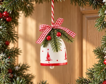 May include: A white bell ornament decorated with red painted pine trees and dots. The bell features a red and white gingham ribbon bow, a red striped hanging cord, and a small sprig of green pine needles with two red berries.