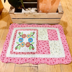 May include: A pink floral quilted mat with a scalloped edge. The center features a white square with embroidered flowers in blue, yellow, and red. The mat is designed for decorative use.