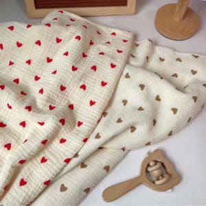 Peut inclure: Couverture de b&eacute;b&eacute; cr&egrave;me avec un motif de c&oelig;urs rouges. Une deuxi&egrave;me couverture avec un motif de c&oelig;urs bruns est visible. Un hochet en bois est &eacute;galement dans l'image. Les couvertures sont en tissu doux et textur&eacute;.
