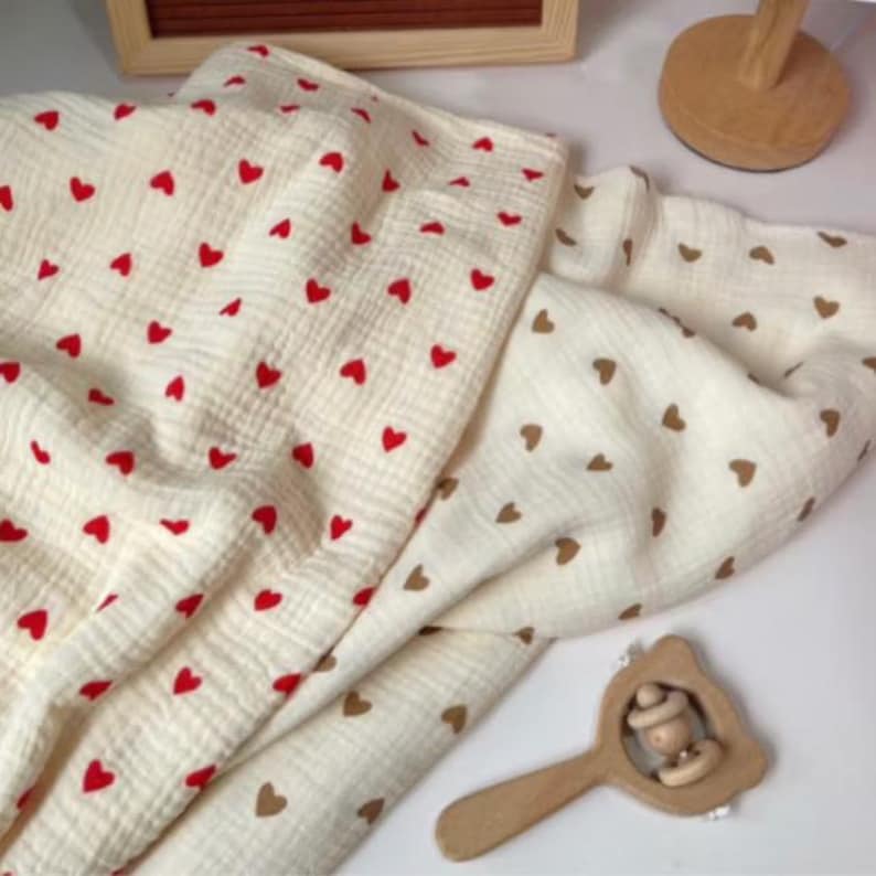 Peut inclure: Couverture de b&eacute;b&eacute; cr&egrave;me avec un motif de c&oelig;urs rouges. Une deuxi&egrave;me couverture avec un motif de c&oelig;urs bruns est visible. Un hochet en bois est &eacute;galement dans l'image. Les couvertures sont en tissu doux et textur&eacute;.