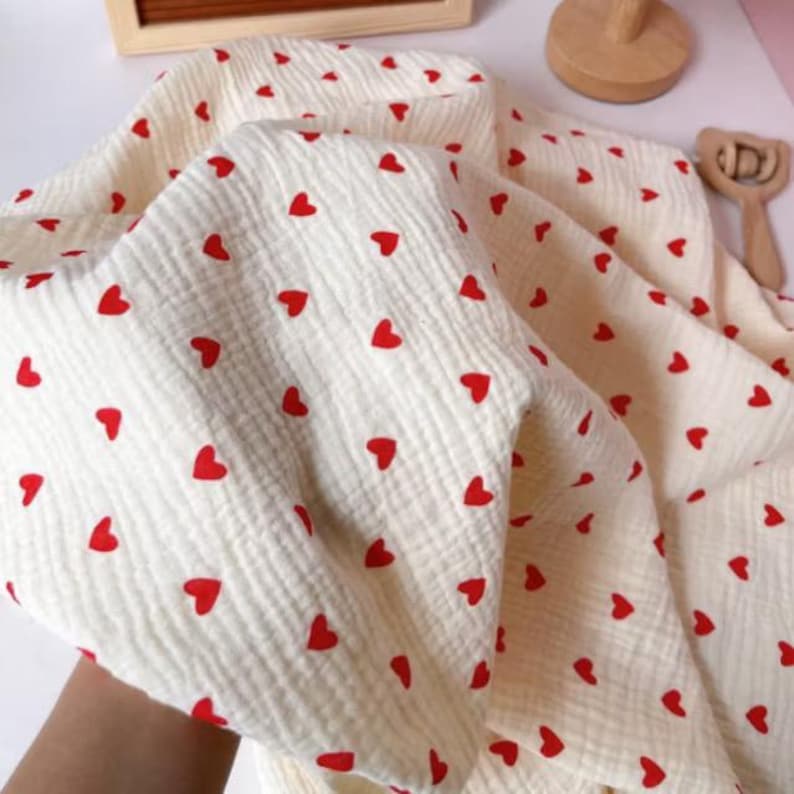 Peut inclure: Tissu en mousseline de couleur cr&egrave;me orn&eacute; de petits c&oelig;urs rouges. Le tissu pr&eacute;sente une texture froiss&eacute;e. L'image montre le tissu pli&eacute;, sugg&eacute;rant une utilisation pour des couvertures pour b&eacute;b&eacute;s ou des articles similaires. Le tissu est doux.