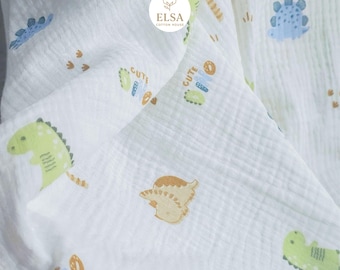 May include: White muslin baby blanket with a repeating pattern of cartoon dinosaurs in green, blue, and tan. The word "CUTE" is printed on the blanket. The ELSA COTTON HOUSE logo is in the upper right corner.