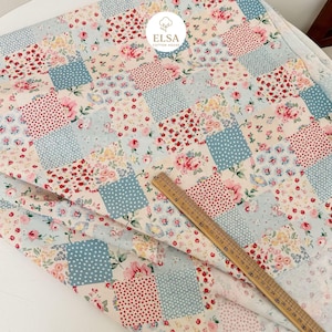 May include: Patchwork fabric with a floral pattern in red, pink, and yellow on a light blue and cream background. The fabric is arranged in a grid of squares, some with polka dots. A wooden ruler is placed on the fabric.