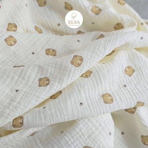 May include: Cream-colored muslin fabric with a repeating pattern of brown teddy bear faces and small paw prints. The fabric is soft and textured, with visible folds and wrinkles. The logo "ELSA COTTON HOUSE" is visible in the upper right corner.