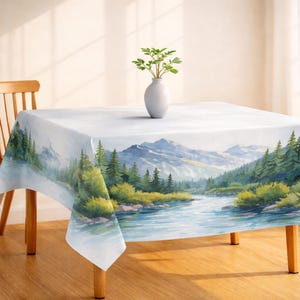 Puede incluir: Un mantel cuadrado con un paisaje de acuarela de un río que fluye a través de un valle con montañas al fondo. La paleta de colores incluye azules, verdes y blancos. Un jarrón blanco con vegetación está sobre la mesa.