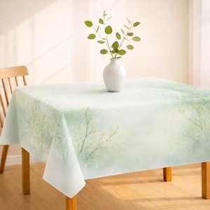 May include: A rectangular tablecloth with a soft green and white watercolor design featuring delicate branches and leaves. A white vase with green sprigs sits on the table. The table is made of wood.