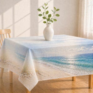 May include: A square tablecloth featuring a beach scene with a blue ocean, white sand, and a light blue sky. A white vase with green sprigs sits on the table. The table is made of wood.