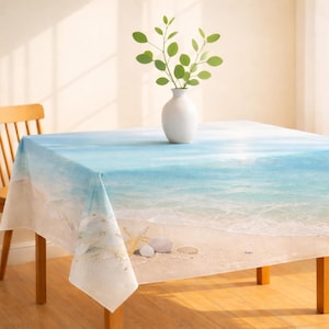 May include: A rectangular tablecloth featuring a beach scene with a blue ocean, white waves, and sandy shore. The design includes seashells and starfish. A white vase with green leaves sits on the table. The table is made of wood.