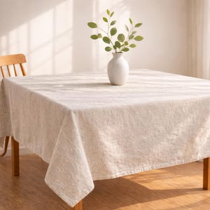 May include: A beige linen tablecloth draped over a wooden table. A white vase with green leafy branches sits in the center. The table is set in a well-lit room, with a wooden chair visible in the background.