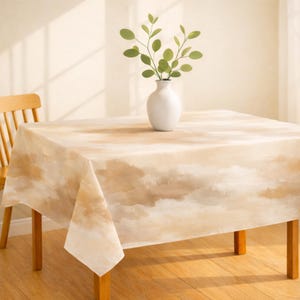 May include: A square tablecloth with a beige and brown watercolor-style design. A white vase with green leafy branches sits on the table. The table has wooden legs and is next to a wooden chair.