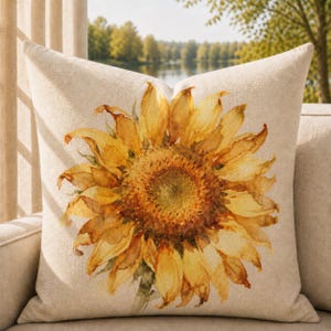 May include: A square throw pillow with a light beige, textured fabric cover. The center of the pillow features a large, detailed watercolor-style illustration of a single sunflower with vibrant yellow and golden-brown petals and a dark center.