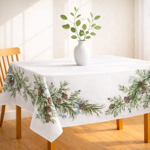 May include: White rectangular tablecloth with a festive winter design. The edges feature a garland of green pine needles, eucalyptus leaves, and brown pinecones. A white vase with green branches sits on the table.