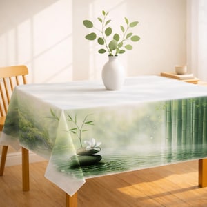 May include: A rectangular tablecloth with a serene bamboo forest and water lily design. The white tablecloth features a green and white landscape with bamboo stalks, water, and a stack of stones with a white flower. A white vase with green leaves sits on the table.