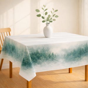 May include: A white tablecloth with a forest scene in shades of green and blue. A white vase with green sprigs sits on the table. The table has wooden legs and a wooden chair is visible in the background.