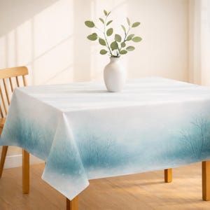 May include: A white tablecloth with a blue and teal watercolor tree design. A white vase with green leafy branches sits on the table. The table is made of wood and has a light brown color.