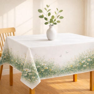 May include: A white square tablecloth with a floral border featuring daisies and green grass. A white vase with green sprigs sits on the table. The tablecloth is draped over a wooden table.