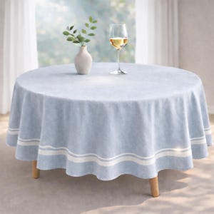 May include: A round, light blue tablecloth with white stripes near the edge. A glass of white wine, a small white vase with greenery, and a wooden-legged table are on the table. The tablecloth is made of a soft fabric.