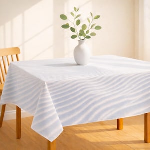 May include: A white tablecloth with a blue wave pattern covers a wooden table. A white vase with green leafy branches sits in the center. A wooden chair is partially visible in the background. The scene is bathed in natural light.