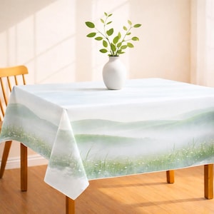 May include: A square tablecloth with a landscape design featuring rolling green hills and a misty atmosphere. A white vase with green sprigs sits on the table. The table is made of wood and has a light brown color.