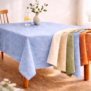 May include: A light blue linen tablecloth covers a wooden table, with a variety of colored linen napkins draped over the edge. A glass of water with a lemon slice and a small vase with greenery are on the table.