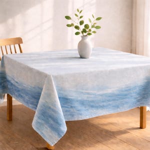 May include: A square tablecloth with a watercolor-style design in shades of blue and white, resembling ocean waves. A white vase with green sprigs sits on the table. The table is made of wood.