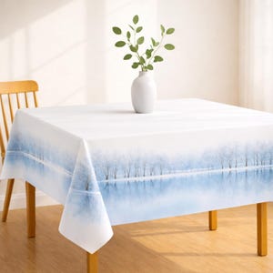 May include: A white rectangular tablecloth with a winter landscape design. The design features a serene scene of a snow-covered forest reflected in a calm lake. A white vase with green sprigs sits on the table. The table is supported by wooden legs.
