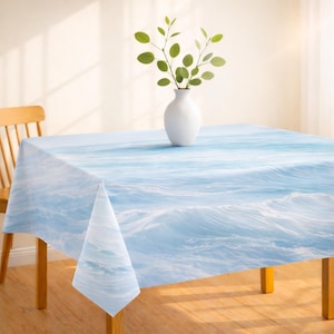 May include: A square tablecloth with a blue and white ocean wave design. A white vase with green leafy branches sits on the table. The table is made of wood and has a light brown color.
