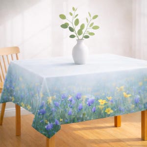 May include: A square tablecloth with a floral design featuring purple and yellow flowers in a field of green grass. A white vase with green leaves sits on the table. The table is made of wood.
