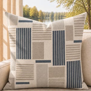 May include: A square throw pillow featuring a geometric pattern of rectangular blocks in shades of beige, cream, and dark blue. The design consists of alternating sections of vertical dark blue stripes and horizontal beige stripes on a cream background.