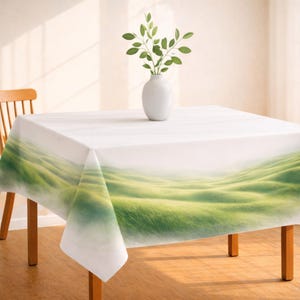 May include: A white square tablecloth with a green and yellow landscape design. The design features rolling hills with a misty effect. A white vase with green leaves sits on the table. The table has wooden legs.