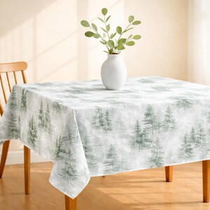 May include: A white tablecloth with a repeating pattern of green pine trees. A white vase with green leafy branches sits on the table. The table is made of wood and has a wooden chair next to it.