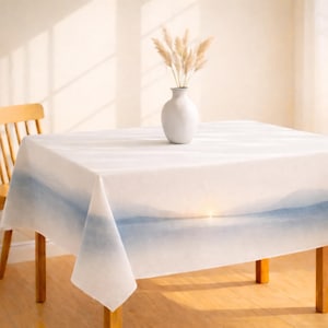 May include: A white tablecloth with a blue watercolor landscape design. A white vase with dried pampas grass sits on the table. The table is made of wood and has a wooden chair next to it.