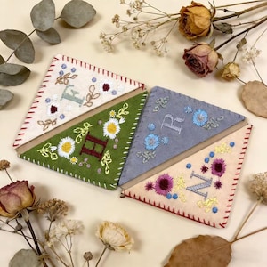 Puede incluir: Cuatro marcadores de fieltro hechos a mano en forma triangular, cada uno con una inicial bordada y un diseño floral diferente. Los colores incluyen crema, verde, gris y beige, con costuras rojas en los bordes. Las letras son E, H, R y N.