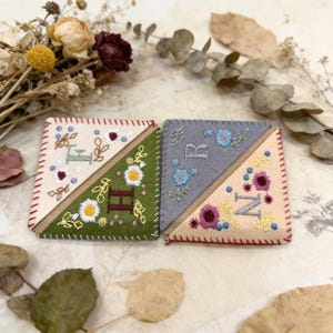 Peut inclure: Deux marque-pages artisanaux en feutre avec des motifs floraux brodés et des lettres initiales. L'un des marque-pages présente les lettres "E" et "H", tandis que l'autre affiche "R" et "N". Chaque marque-page a un code couleur différent, avec une bordure cousue.