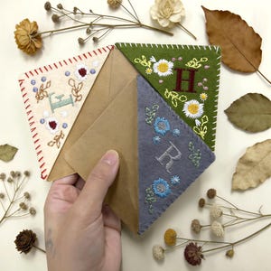 May include: Hand holding a brown paper envelope with three embroidered felt corner bookmarks. The bookmarks are cream, green, and gray, each with floral embroidery and a single initial. The cream bookmark has red stitching.