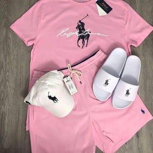 May include: A pink t-shirt, shorts, cap, and slides set. The t-shirt features a black logo and the name "Ralph Lauren" in white script. The white cap and slides also have the black logo. The shorts have a drawstring and the same logo.