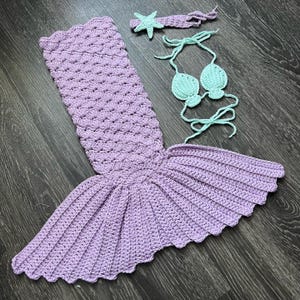 May include: A crocheted mermaid costume set in shades of lavender and mint green. The set includes a mermaid tail, a bikini top, and a starfish headband. The tail is a light purple color with a scalloped edge.