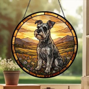 May include: Round stained glass window art featuring a Schnauzer dog sitting in a field with a mountain backdrop. The artwork has a black border and is suspended by a chain. The background is a warm sunset color.