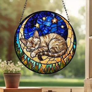 Könnte beinhalten: Rundes Buntglasfenster mit einer schlafenden Katze auf einem Mond vor einem Sternenhimmel. Die Katze ist braun und beige, der Mond ist gelb und der Himmel ist blau. Das Fenster hängt an einer Kette.
