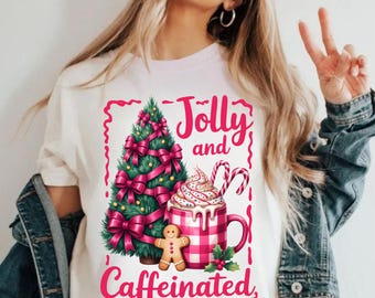 May include: White t-shirt with a festive graphic. The design features a decorated Christmas tree with pink bows, a mug of whipped cream, candy canes, and a gingerbread man. The words "Jolly and Caffeinated" are written in pink.
