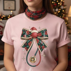 May include: A pale pink t-shirt with a festive design. The graphic shows a bow with red flowers and green foliage, a Santa hat, and a stethoscope. The stethoscope is red and green with a gold-coloured chest piece.