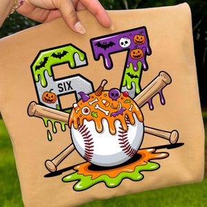 May include: Tan baseball-themed graphic featuring the numbers "67" in green and purple, adorned with Halloween motifs such as bats, pumpkins, and a skull. A baseball with orange dripping icing and crossed bats complete the design. The word "SIX" is also visible.